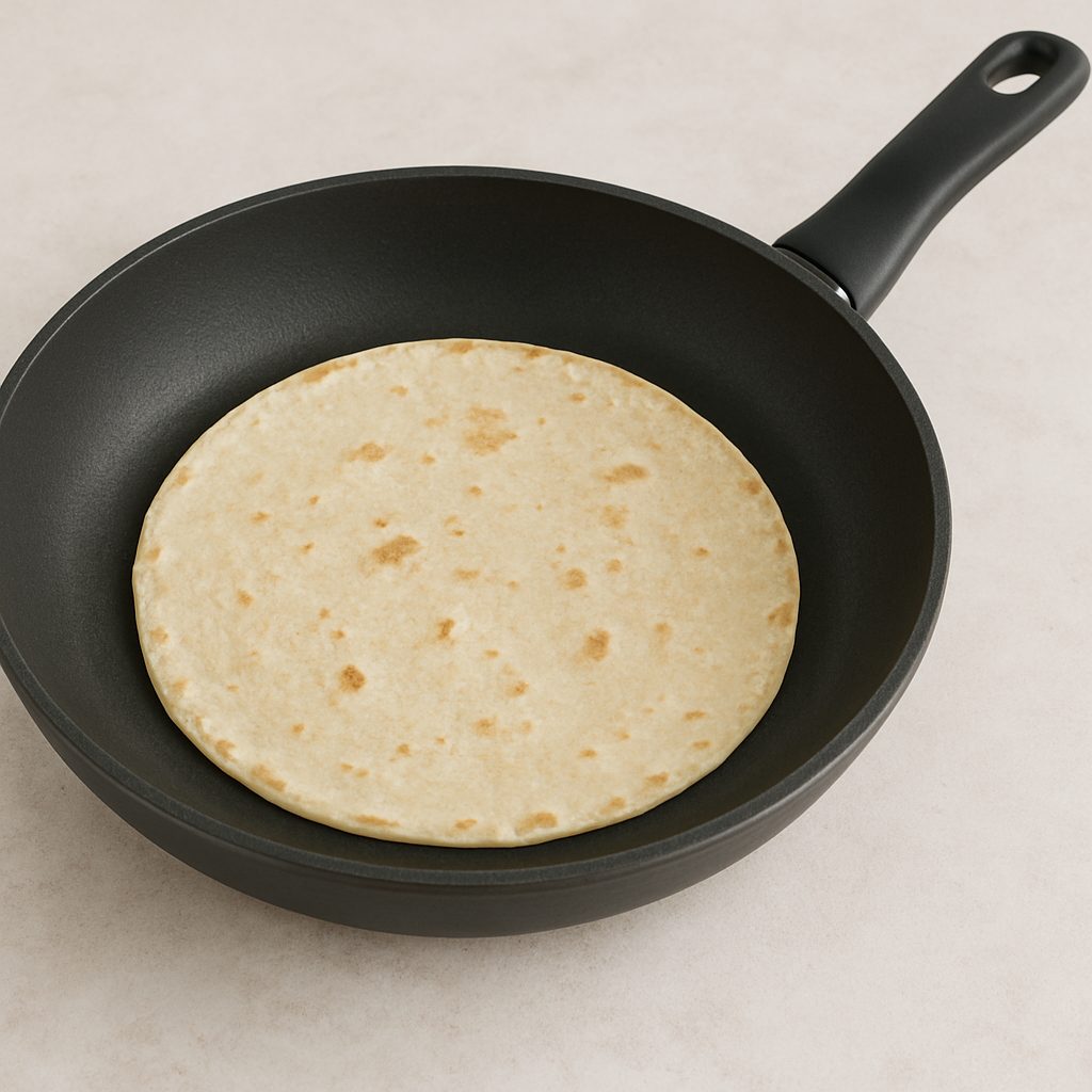 "Tortilla de maíz calentándose en una sartén negra sobre una superficie clara, con ligeras marcas doradas."
